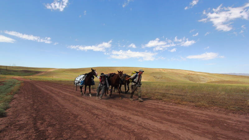 Wide shot of two men and three horses walking down a dirt road in Kazakhstan.