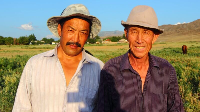 Two Kazakhstan men smile for the camera in the sunshine, wearing wide hats.