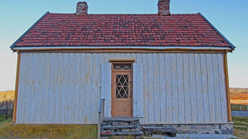A frontside shot of a white shiplap constructed house, with a red roof and brown door.