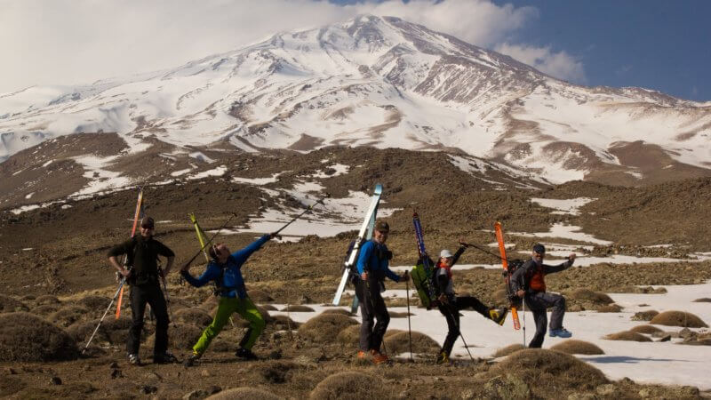 Photo of Mount Damavand in distance with five team members in foreground wearing skis on their backs and throwing funny poses.