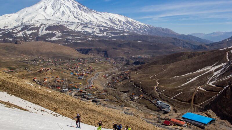 Photo of Mount Damavand in background and five ski mountaineers on a nearby peak in foreground. In the middle is a very wide valley with a few dozen houses.