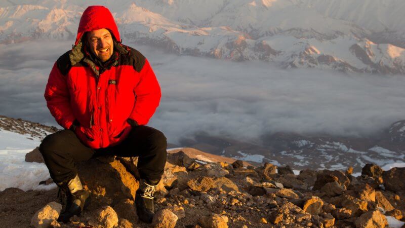 Happy skier sat on a rock high on the slopes of Damavand. Below is a cloud-filled valley.