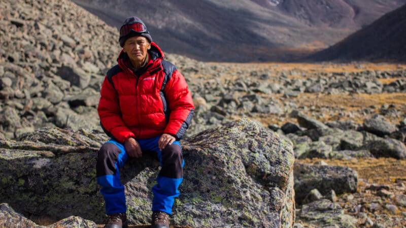 A Tuvan mountaineer wearing blue trousers and red jacket sat on a rock posing for the camera.
