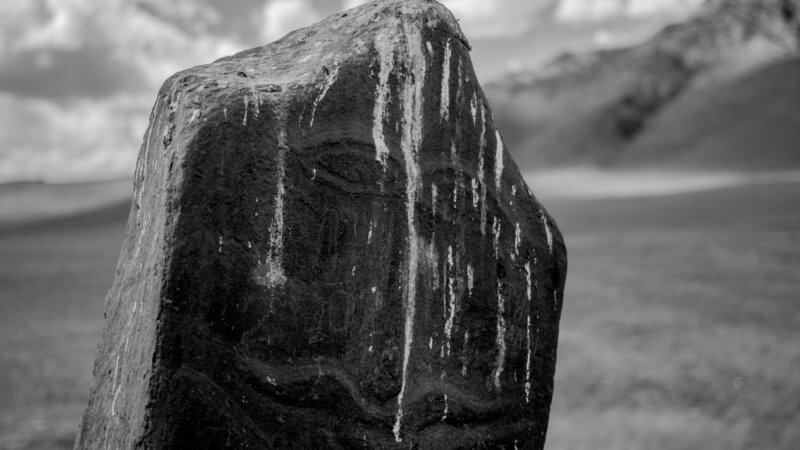 Black and white photo of a Scythian-era standing deer stone in Tuva with a moustached faced carve on it.