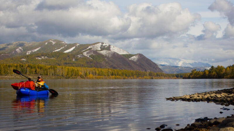 A man paddling a blue packraft descends the Yenisei River with snow-covered mountains behind.