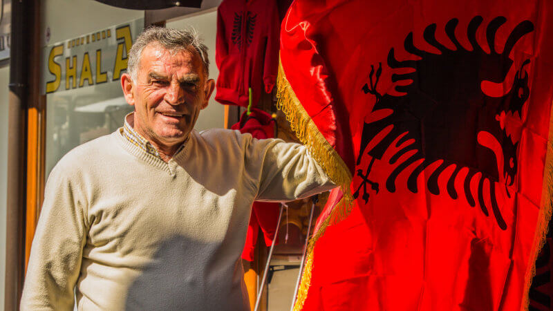 Man in white sweater poses for camera holding up an Albanian flag.