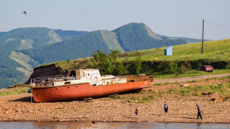 Three boys in the distance play next to an abandoned red hulled fishing vessel that's aground.