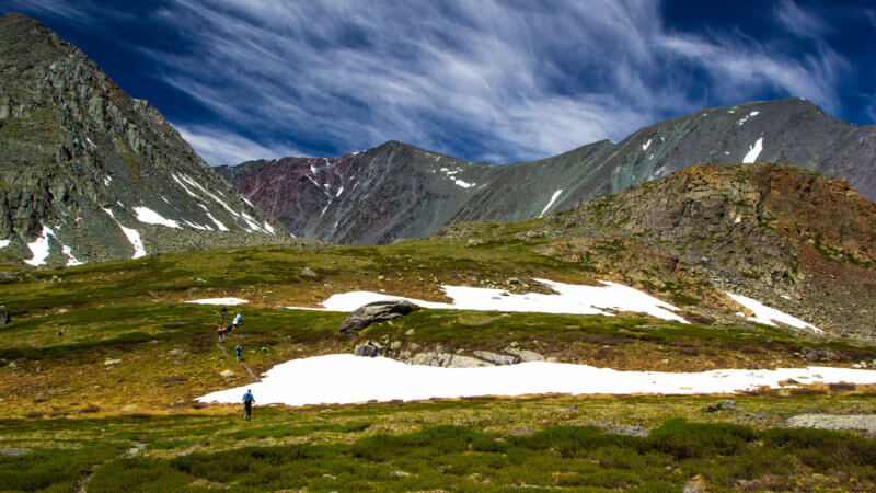 A group of walkers ascend over grassy hills and beneath blue skies.