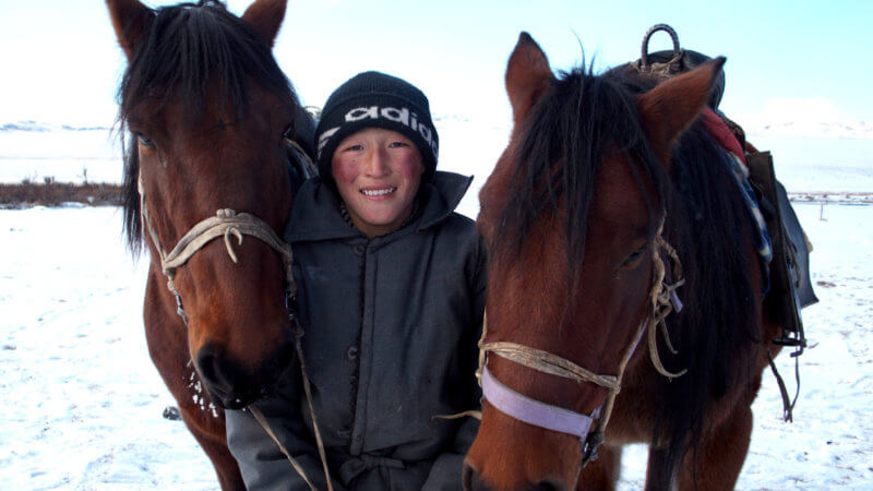 A Kazakh boy in winter grins while holding two horses either side of him.