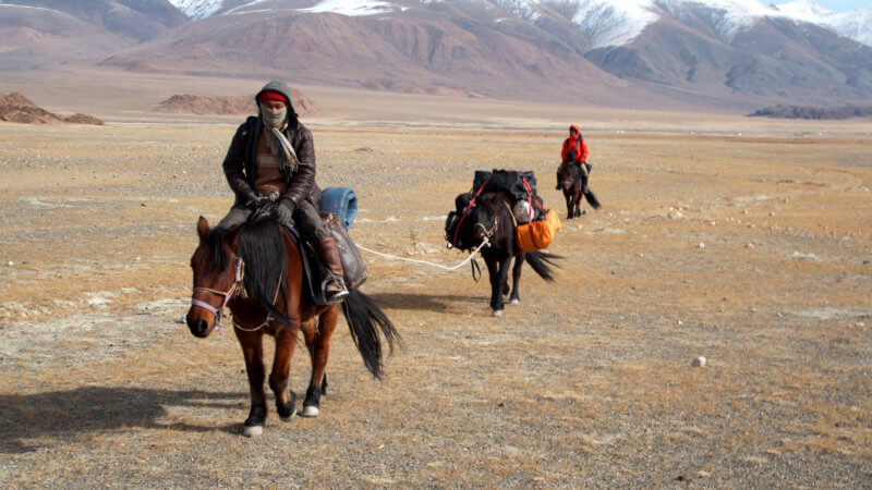 Three horses and two guys loaded with equipment, riding across a flat Mongolian steppe.