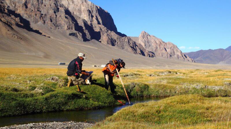 Two men, one holding a fishing net, lean into a small river to catch fish.