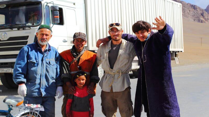 Two Tajik truck drivers stand beside three others for a photo and wave.